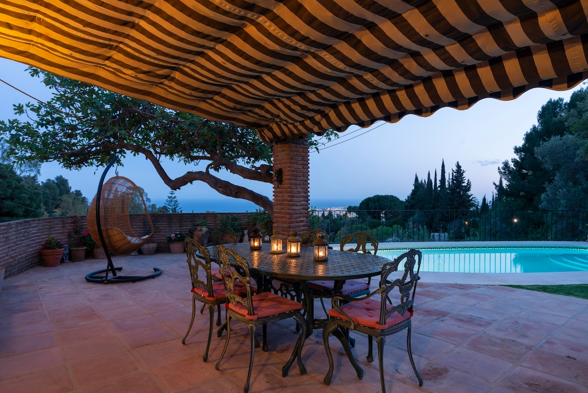 An outdoor dining area with a striped awning features a table surrounded by elegant chairs. Lanterns are placed on the table, casting a soft glow. In the background, a swimming pool reflects the evening sky, bordered by trees and plants.