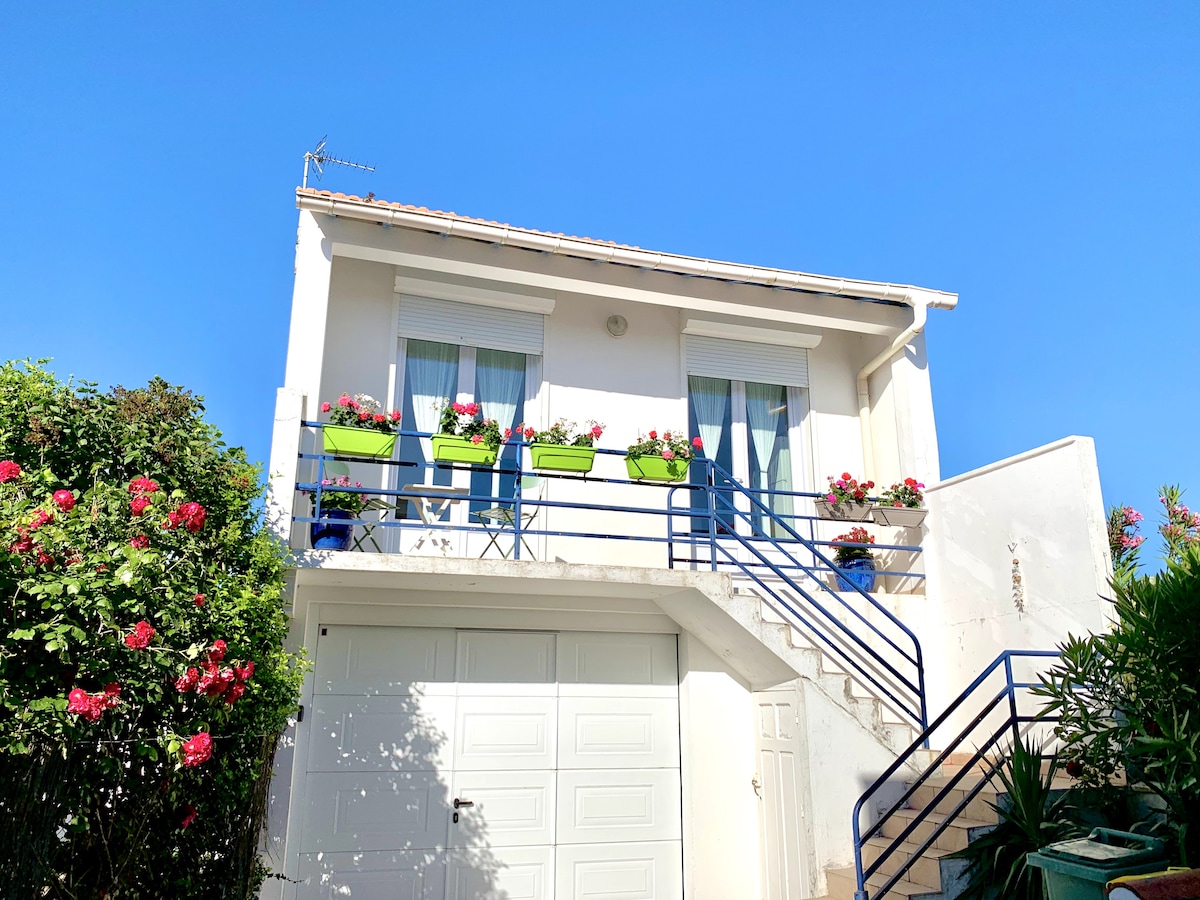 A charming two-story house is set against a clear blue sky. Flower boxes adorn the windows, filled with vibrant plants. A flight of stairs leads up to the entrance, framed by lush greenery and flowering shrubs.