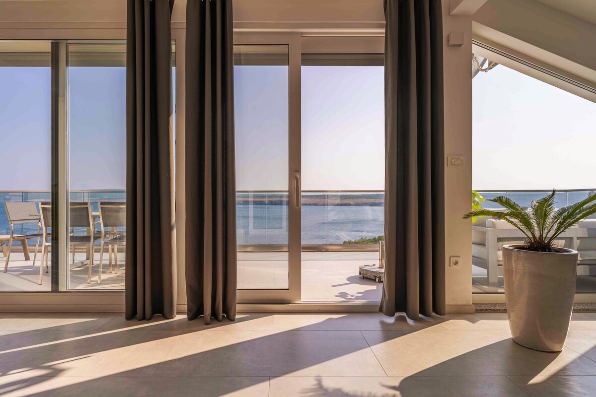 Large glass doors provide a clear view of the sea and balcony. Neutral-toned curtains frame the entrance, while a potted plant adds a touch of greenery. Natural light fills the space, enhancing the calm ambiance of the room.