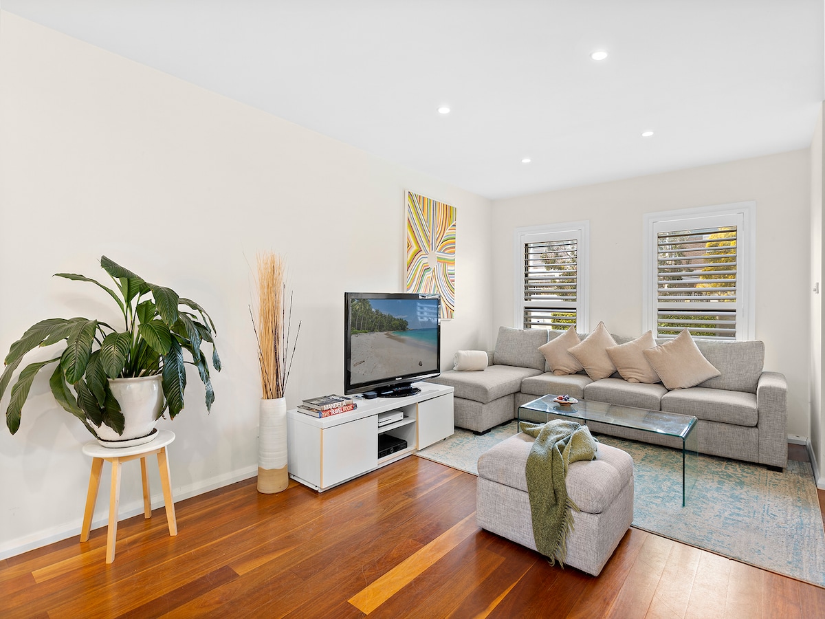 A spacious living area is featured with a large sectional sofa adorned with decorative cushions. A coffee table sits atop a light-colored rug, and a television is mounted on a sleek media console. Natural light filters through the window, complemented by potted greenery.