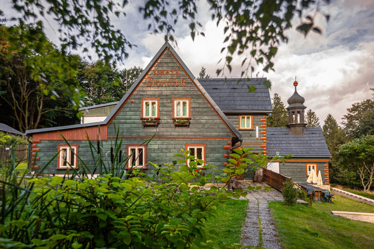 The exterior of a charming chalet is showcased, featuring a wooden structure with a pitched roof and unique architectural details. Lush greenery surrounds the building, and flower boxes enhance the facade. A gravel path leads to the entrance, providing access to the serene environment.