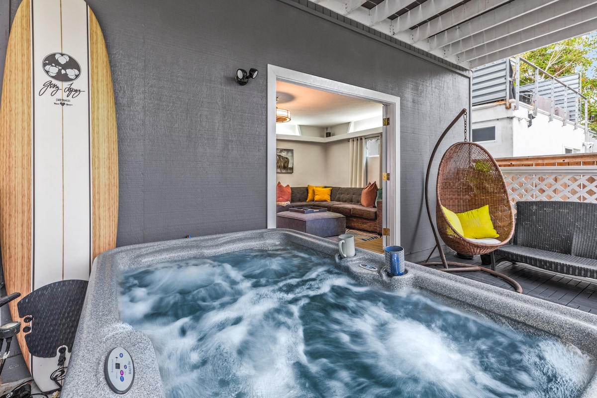 A hot tub bubbles invitingly on a patio, surrounded by gray walls. A stylish surfboard leans against the wall. The interior space is partially visible through an open door, showcasing a comfortable seating area with a bright yellow cushion.