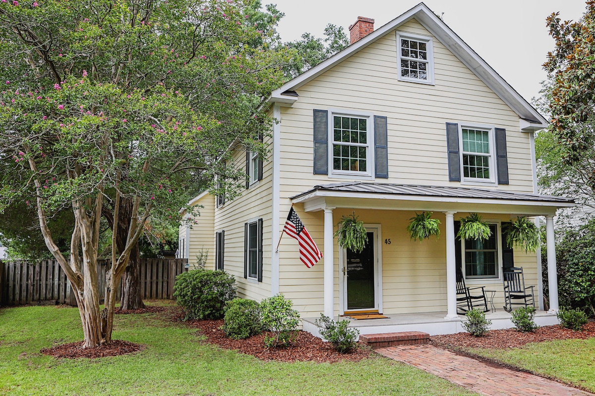 A charming historic home is framed by lush greenery and features a front porch with a rocking chair. The exterior is painted in soft yellow with dark shutters. An American flag hangs beside the door, enhancing the inviting entrance.