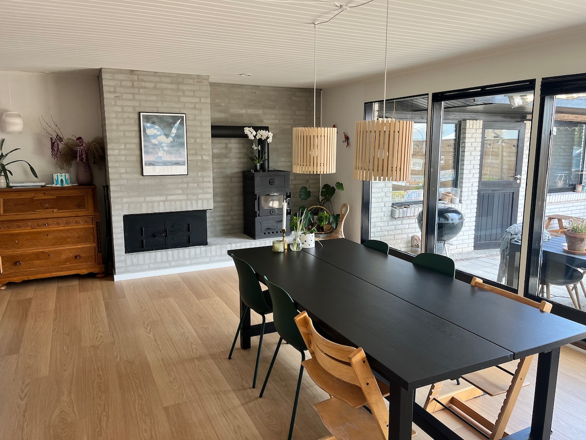 A modern dining area features a long black table surrounded by wooden and green chairs. A stone fireplace is positioned against the wall, complemented by natural light streaming in through large glass doors, which open to an outdoor space.