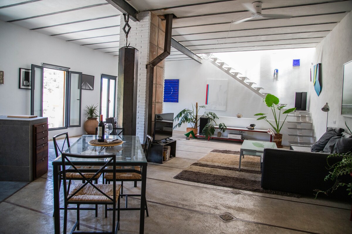 An open living space features a dining area with a glass table and black chairs. Natural light floods through large windows, highlighting a cozy seating area with a dark sofa. A staircase leads to the upper level, accented by contemporary artwork and indoor plants.