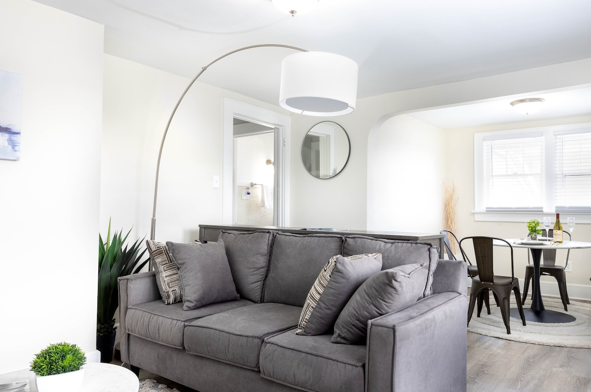 A spacious living area features a gray sofa adorned with decorative pillows. A curved floor lamp stands beside the sofa. In the background, a round dining table with black chairs is visible, along with a soft area rug that adds to the inviting ambiance.