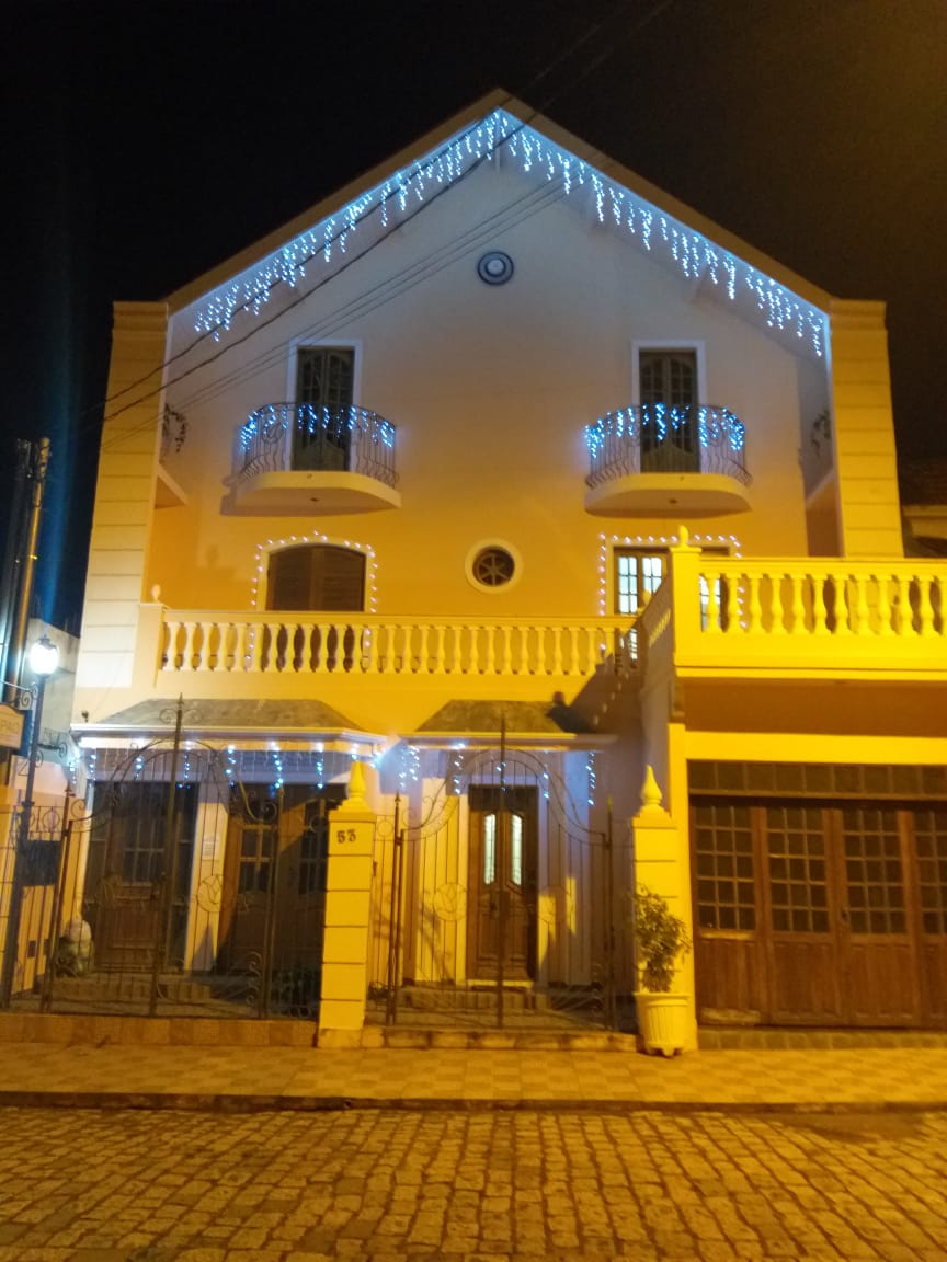 The exterior of the house is adorned with soft blue lights, highlighting the elegant balconies and architectural details. The warm glow from the windows enhances the welcoming atmosphere of the entrance, with cobblestone pathways leading to the inviting front door.
