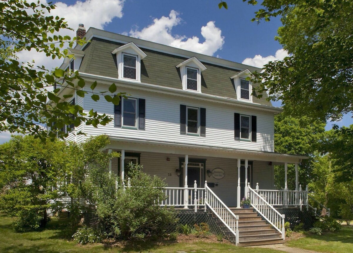 A three-story structure features a classic design with a sloped roof and large front porch. The white exterior is accented by black shutters, and greenery surrounds the home, enhancing the inviting entrance. Bright blue skies and fluffy clouds provide a picturesque backdrop.