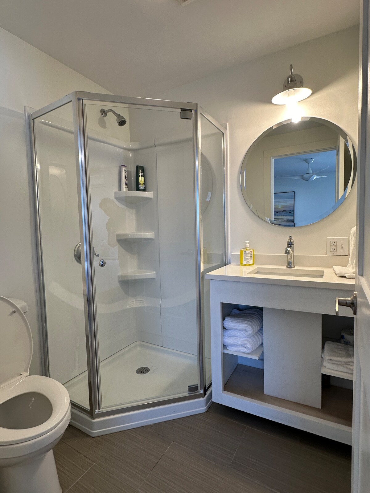 A well-appointed bathroom is presented, featuring a glass-enclosed shower with built-in shelves. A round mirror hangs above the modern sink, which is paired with a compact cabinet storing neatly arranged towels. Ample lighting is provided by a ceiling fixture, enhancing the space.