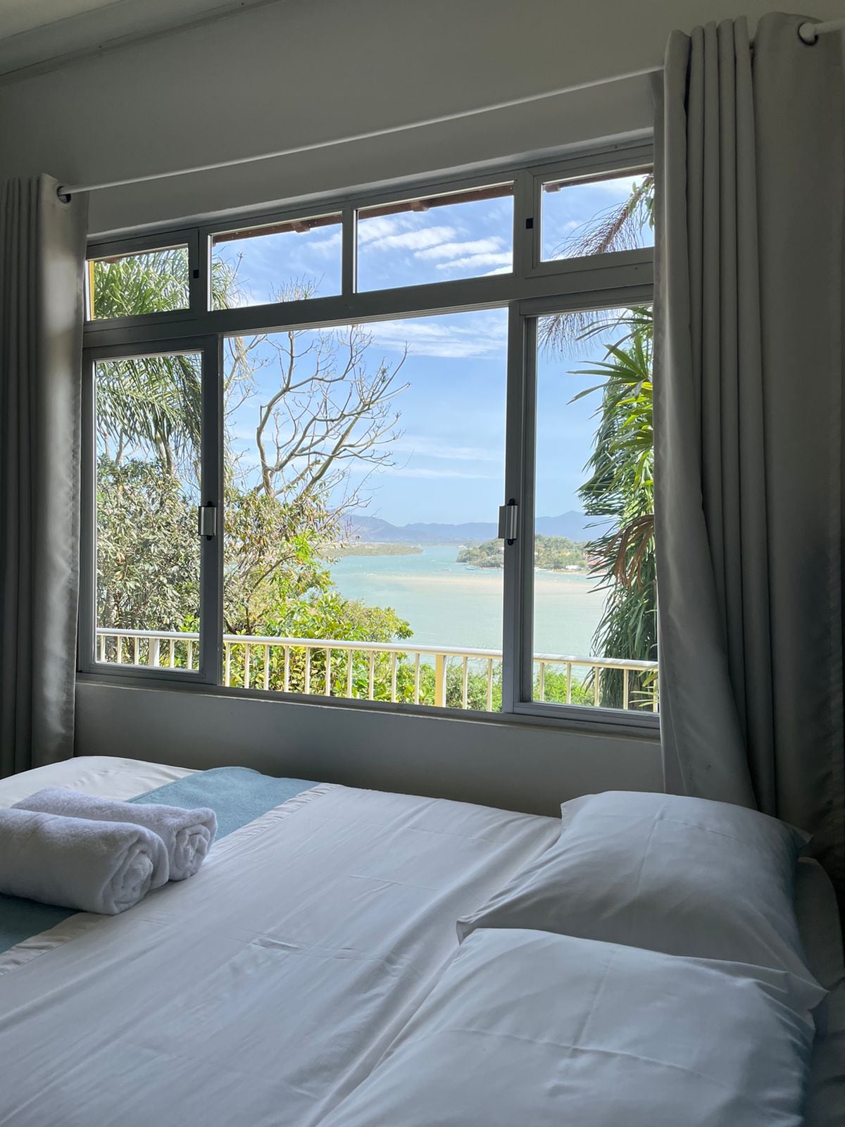 A spacious bed is positioned by a large window, offering a clear view of the beach and surrounding greenery. Soft light enters through the window, enhancing the serene atmosphere of the room.