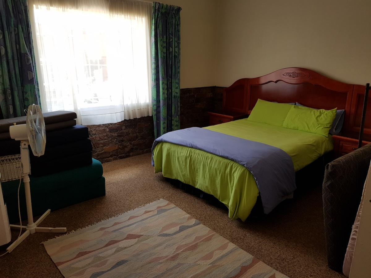 A private bedroom is furnished with a spacious bed covered in vibrant green and blue linens. Soft beige carpet and a decorative rug add warmth to the room. Natural light brightens the space through a large window draped with patterned curtains.
