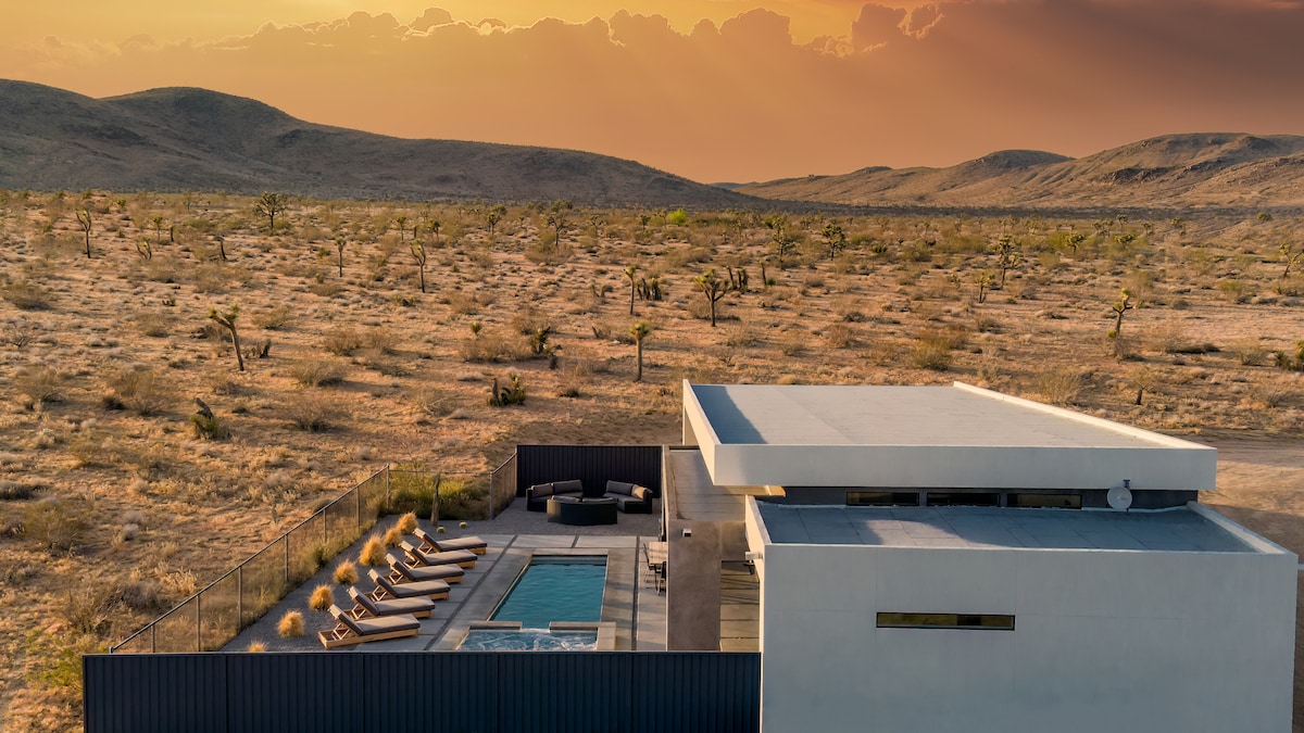 An elevated view reveals a modern home surrounded by the Mojave Desert's arid landscape. The outdoor area features a heated pool, six lounge beds, and a hot tub, while gentle hills and Joshua trees create a serene backdrop illuminated by the setting sun.