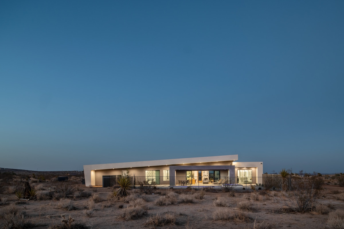 The modern structure showcases a distinctive angled roofline against a twilight sky, set amidst the Mojave Desert landscape. The facade is highlighted by large windows, reflecting the serene surroundings and offering a glimpse of the outdoor living space.