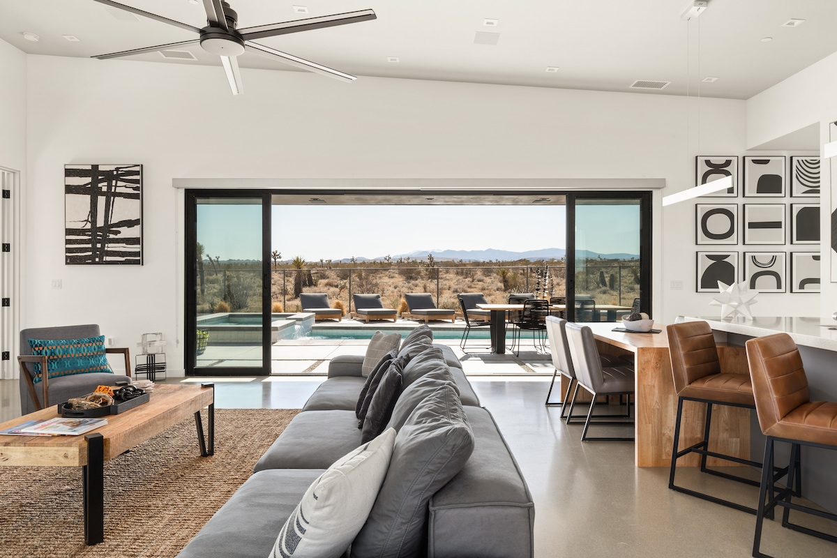 A spacious living area features large sliding glass doors, allowing for abundant natural light and views of the desert landscape. Comfortable seating is arranged around a wooden coffee table. A well-equipped kitchen and dining area can be seen in the background, complementing the modern aesthetic.