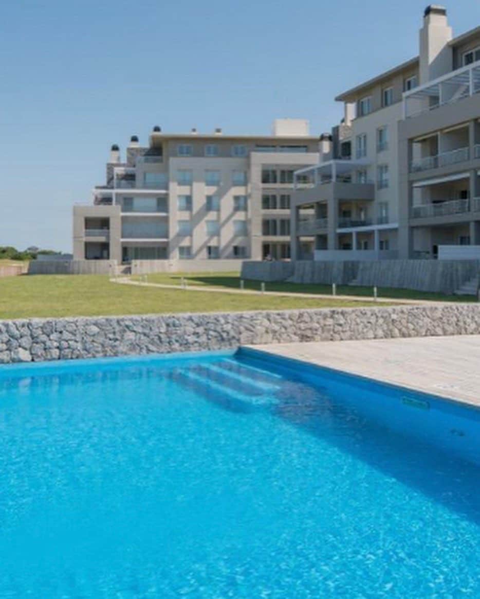 A sparkling blue swimming pool is set against a backdrop of modern apartment buildings. The surrounding area features manicured grassy lawns and a stone retaining wall, offering a serene outdoor space for relaxation. Clear skies enhance the inviting atmosphere of the property.