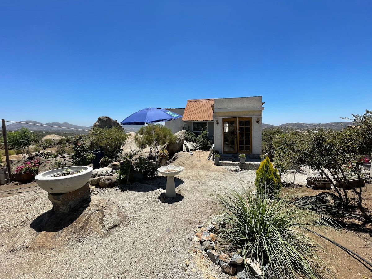 The outdoor space is characterized by a gravel pathway leading to a rustic cabin with a metal roof. An umbrella provides shade over a seating area, while landscaped areas feature plants and stones. A stone water basin is positioned in the foreground.