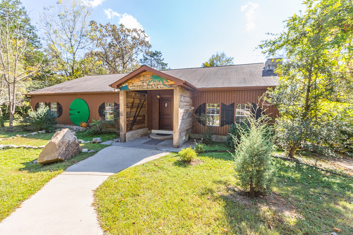 A welcoming exterior view of the lodge showcases a wooden facade with a central entrance framed by stone accents. Lush greenery surrounds the building, complemented by a pathway leading to the door. A round green door adds a whimsical touch to the rustic charm.