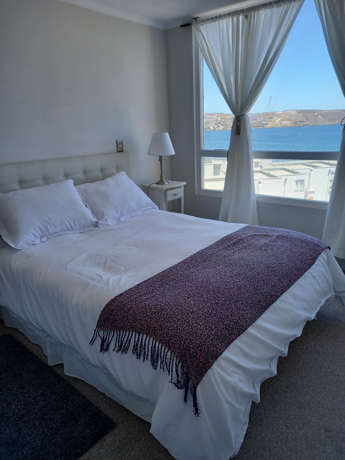 A light-filled bedroom features a spacious bed adorned with a textured throw. A bedside lamp rests on a small table next to the bed. Sheer curtains frame the window, offering a view of the sea and allowing natural light to brighten the room.