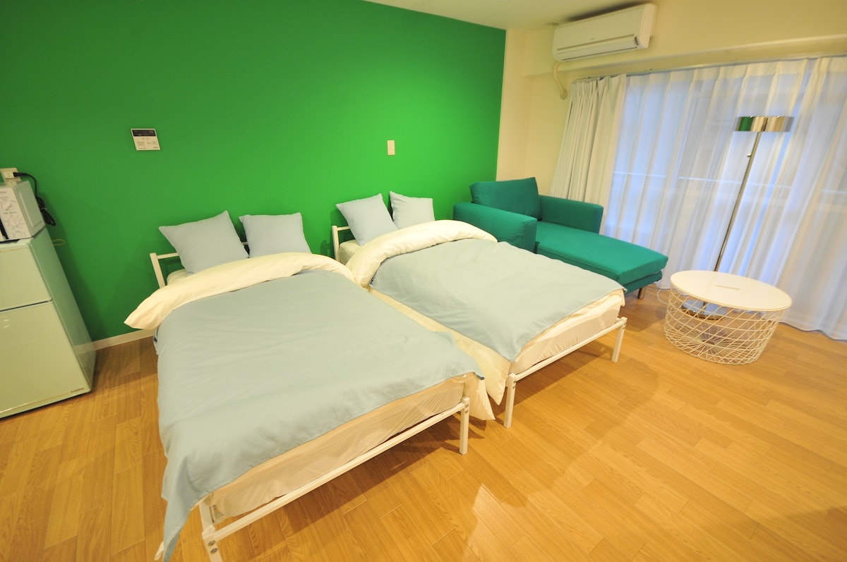 A room features two spacious beds dressed in light blue bedding, placed against a vibrant green wall. A cozy seating area is provided with a green sofa, complemented by a round white table. Natural light enters through a window with sheer curtains.