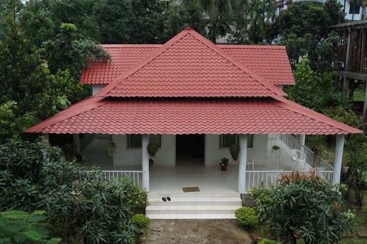 A bungalow features a striking multi-faceted red roof, surrounded by lush greenery and trees. The spacious verandah is supported by white columns, providing an inviting entryway. The light-colored exterior walls complement the vibrant roofing and enhance the overall aesthetic.