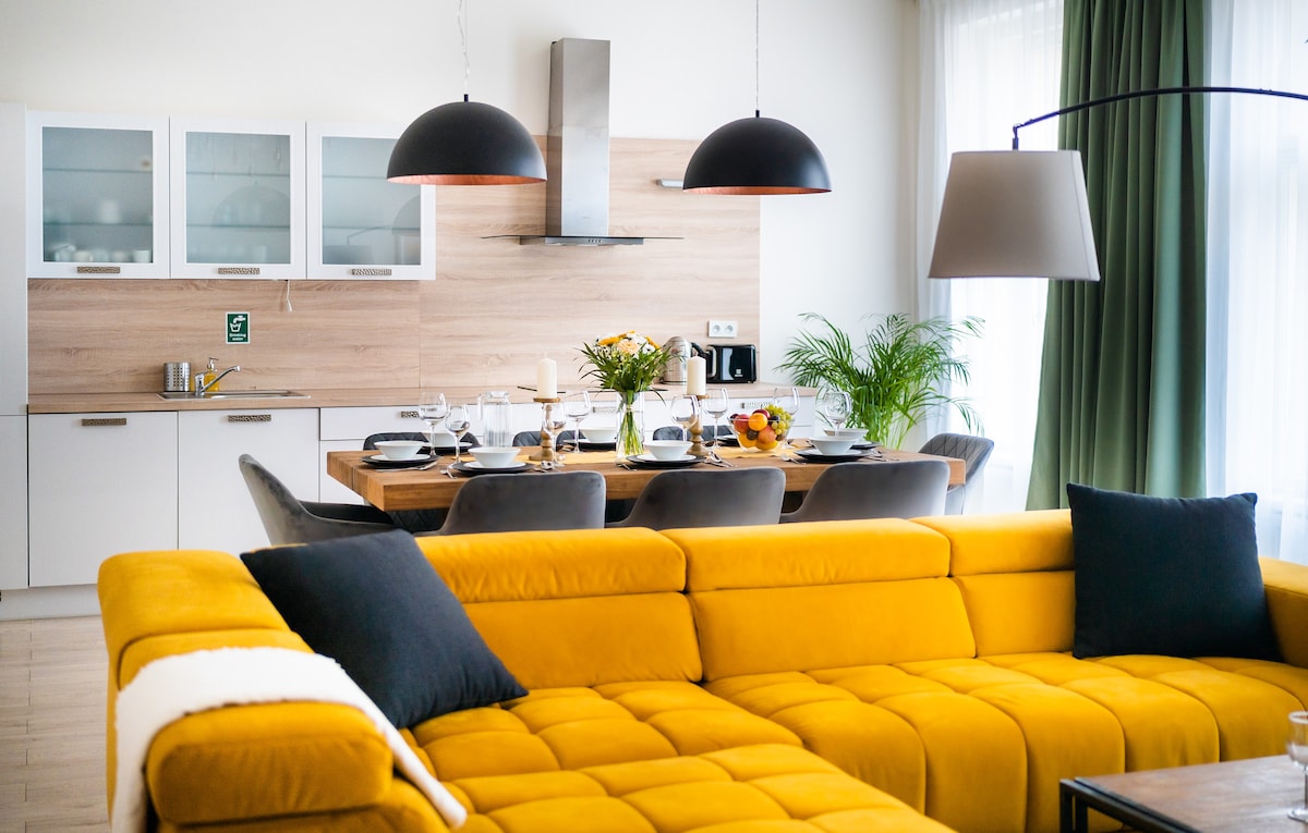 A modern living space showcases a vibrant yellow sectional sofa and a dining table set for multiple guests. Natural light fills the area, highlighting the wooden accents and sleek kitchen appliances. Stylish pendant lights hang above the dining table, adding to the contemporary feel of the apartment.