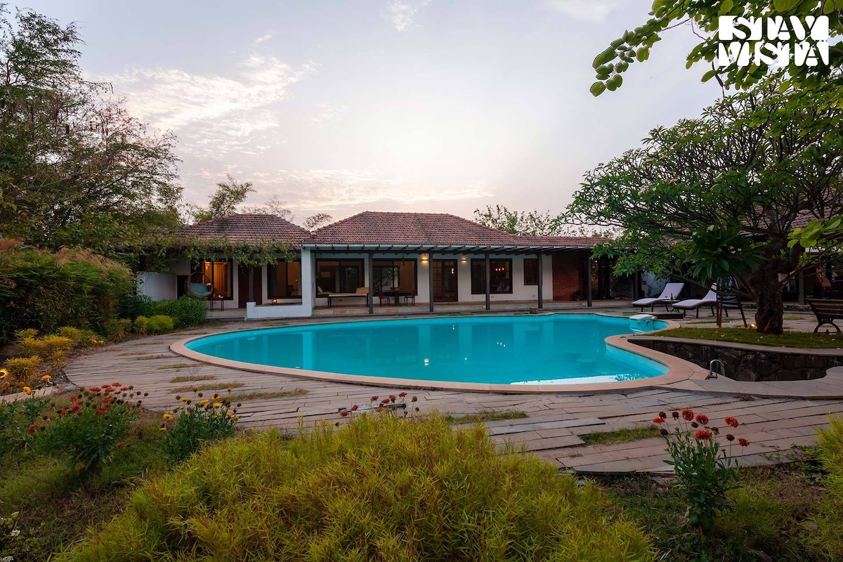 A serene outdoor view features a private swimming pool surrounded by lush landscaping, including colorful flower beds. The villa's modern architecture blends seamlessly with nature, and the sky reflects soft hues as the sun rises, creating a tranquil atmosphere.
