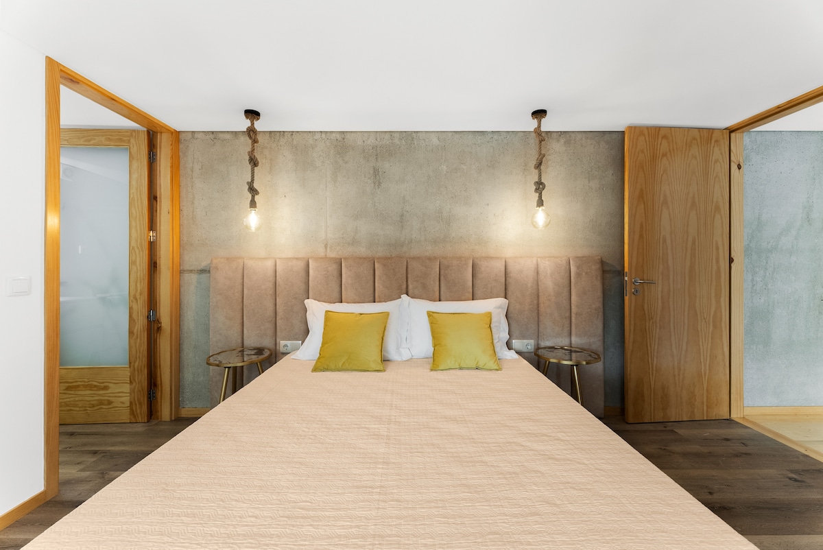 A spacious bedroom features a padded headboard with two decorative pillows. Soft lighting is provided by hanging bulbs on either side. Two wooden doors lead to adjacent rooms, while a neutral-toned bedspread covers the large bed.