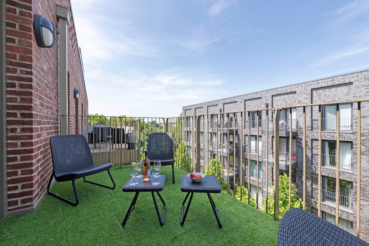 A private balcony is featured with artificial grass, accommodating two modern chairs and a low table. Two glasses and a bottle of wine sit atop the table, with a view of the surrounding buildings and greenery beyond the railing.