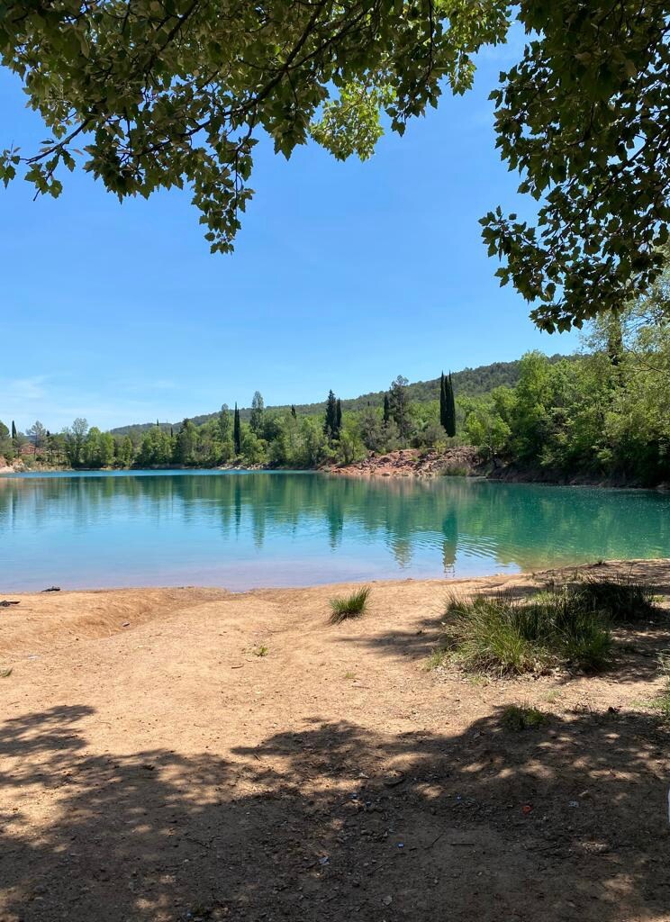 A tranquil natural swimming area features clear turquoise water surrounded by lush greenery and sandy shores. The landscape is accentuated by distant hills and a variety of trees, providing a peaceful outdoor setting ideal for relaxation and leisure activities.