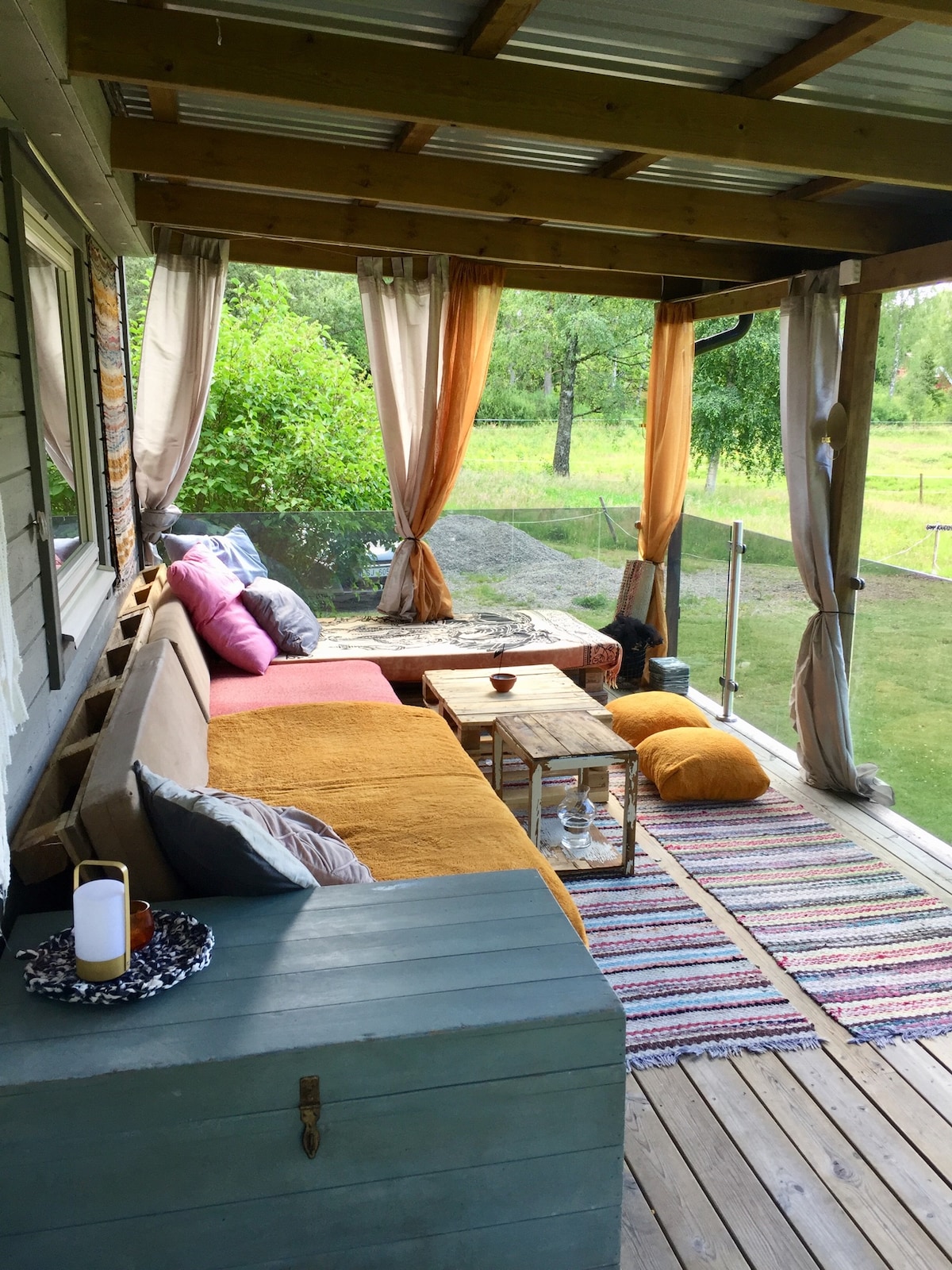 A covered outdoor seating area features a spacious sofa adorned with colorful cushions and throws. A wooden coffee table sits at its center, accompanied by soft floor cushions on a patterned rug. Sheer curtains gently sway in the breeze, allowing views of the lush greenery beyond.