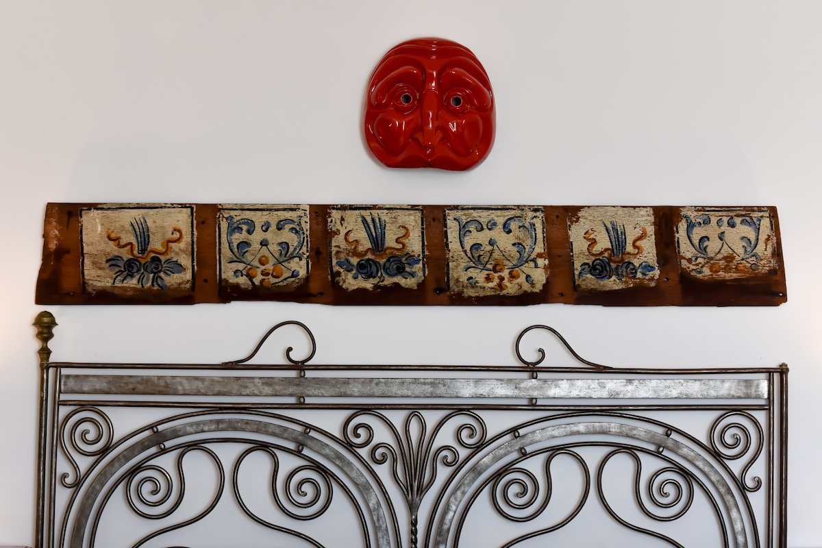 A decorative headboard made of metal is displayed against a white wall. Above the headboard, a horizontal arrangement of five ceramic tiles is visible, featuring floral patterns in blue and yellow. A red ceramic plate is also present but not clearly detailed.