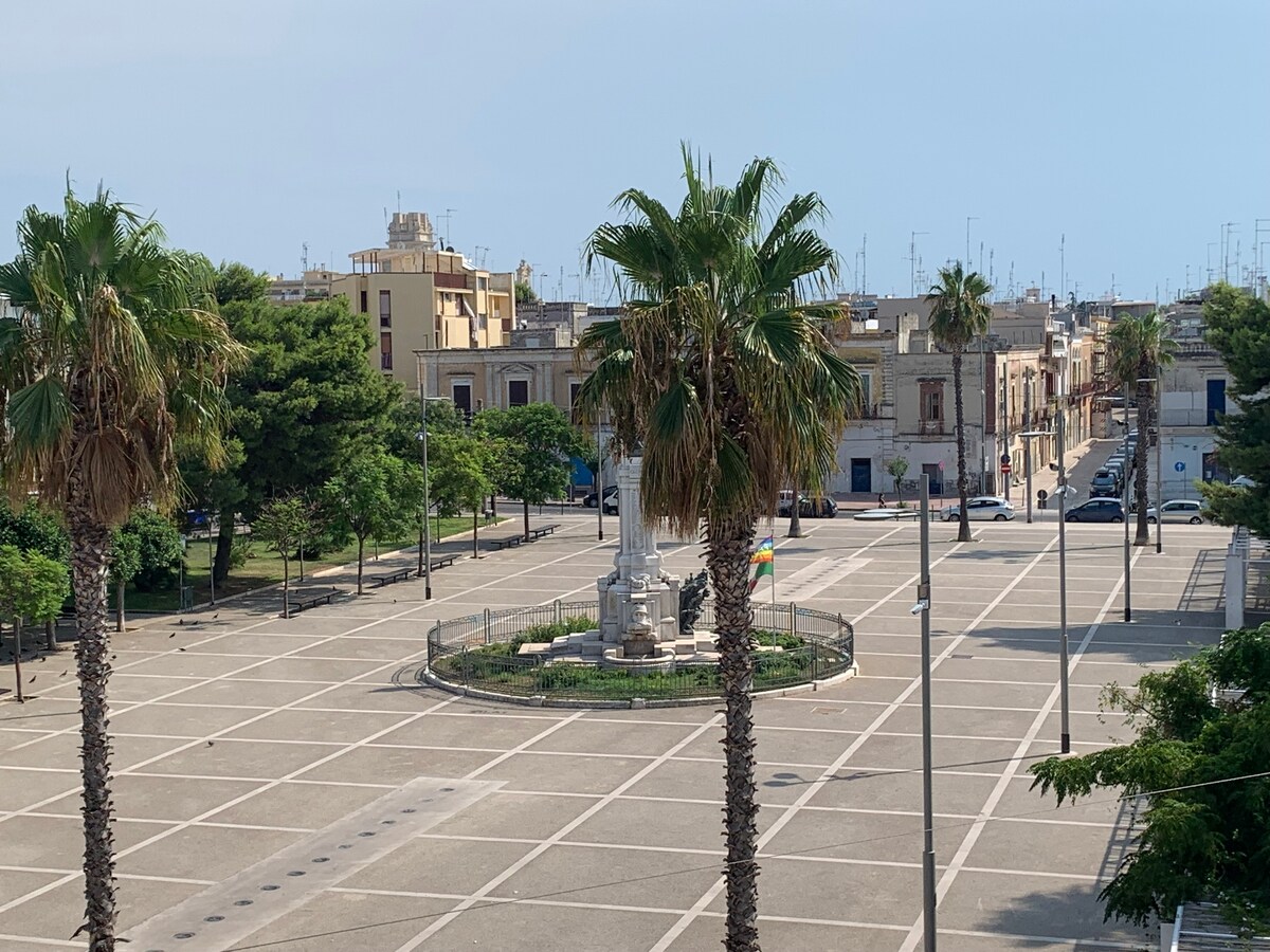 A spacious piazza is shown, featuring palm trees and a central monument. Well-maintained pavements and surrounding buildings provide a charming backdrop. The area is bathed in natural light, with clear blue skies visible overhead.