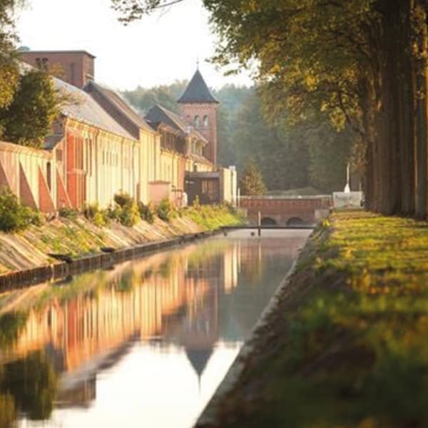 A serene canal reflects the surrounding trees and buildings, creating a calm atmosphere. The pathway lined with greenery leads towards a distant tower, surrounded by a gentle landscape. The warm light enhances the details of the structures along the water's edge.