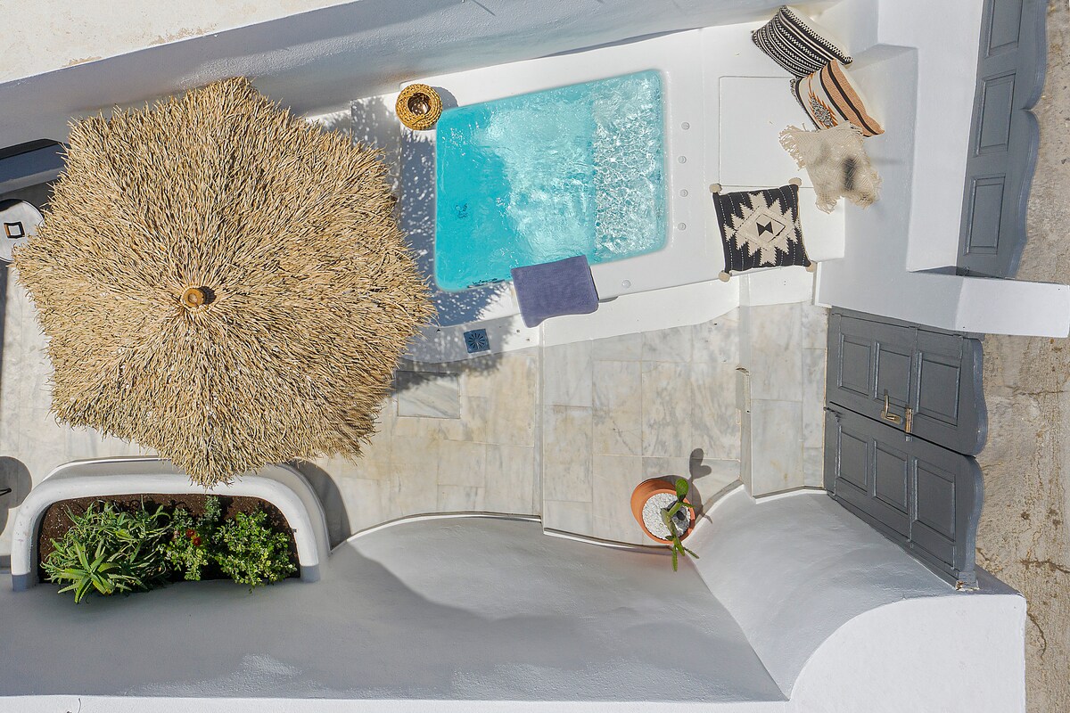 An aerial view of a private courtyard showcases a heatable jacuzzi surrounded by marble flooring. A thatched umbrella provides shade, while potted plants add a touch of greenery. The white walls create a bright atmosphere, highlighting the tranquility of the outdoor space.