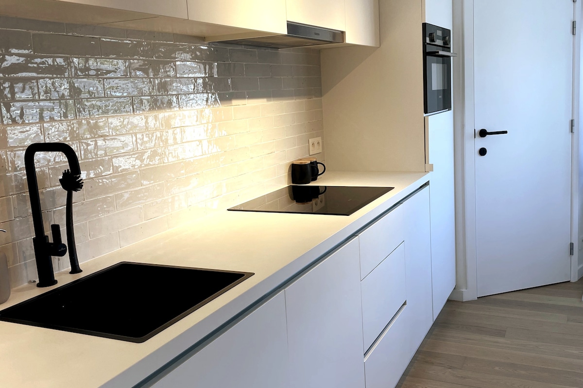 A modern kitchen features sleek white cabinetry and a smooth countertop equipped with a built-in stovetop and sink. A minimalist design is complemented by a stylish black faucet. Light reflects off the glossy backsplash, enhancing the kitchen's clean and contemporary look.