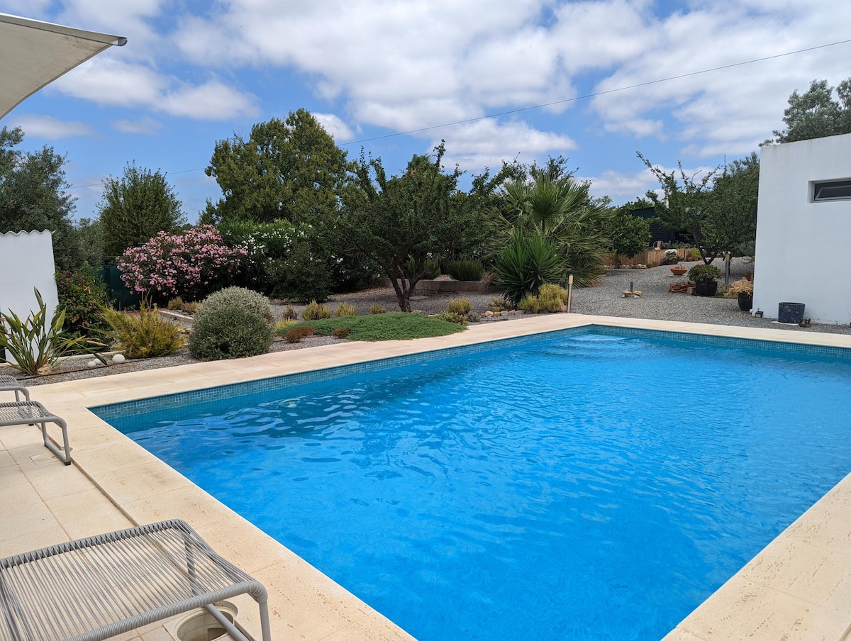 A large swimming pool is framed by neatly landscaped gardens, with various trees, shrubs, and flowering plants visible in the background. The clear blue water of the pool contrasts beautifully with the light-colored stone coping surrounding it.