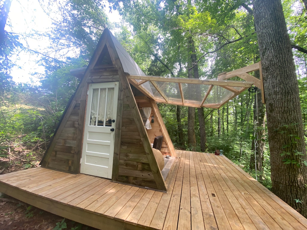 An A-frame cabin is situated on a wooden deck, surrounded by lush greenery. The cabin features a door with a grid-style window, while a transparent canopy extends over the deck, providing a shaded area for relaxation amidst the trees.