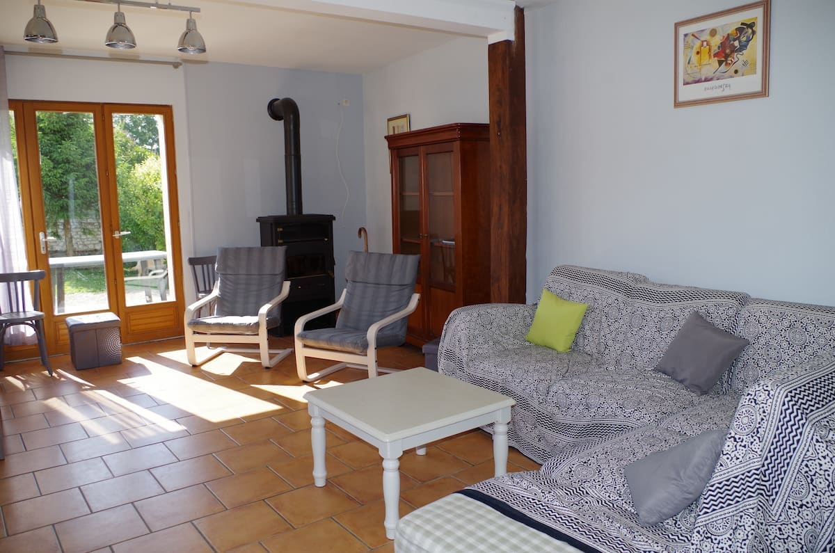 A spacious living area is highlighted by natural light streaming through large glass doors. Comfortable seating is arranged, including a patterned sofa and two armchairs. A small coffee table sits center stage, while a wooden cabinet and a charming wood stove complete the inviting space.