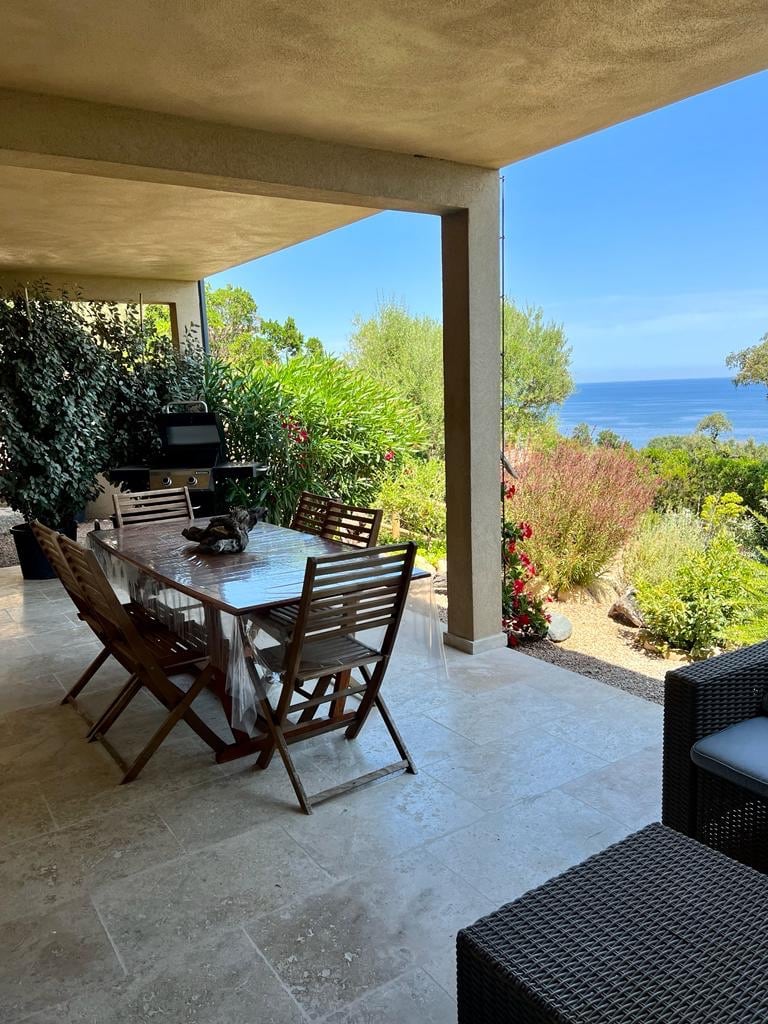 A covered terrace features a large dining table surrounded by wooden chairs. Lush greenery and flowering plants frame the area, with the ocean visible in the background. The space is complemented by comfortable seating on one side, enhancing the inviting atmosphere.