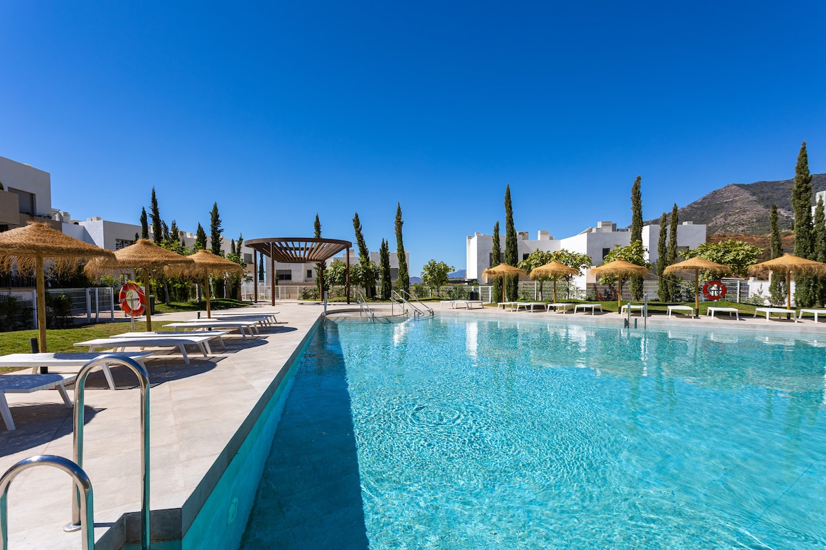A large communal swimming pool is surrounded by a tranquil setting, featuring several sun loungers and umbrellas. Lush greenery lines the area, with tall cypress trees adding to the serene atmosphere. The clear blue water reflects the sunny sky, creating an inviting outdoor retreat.