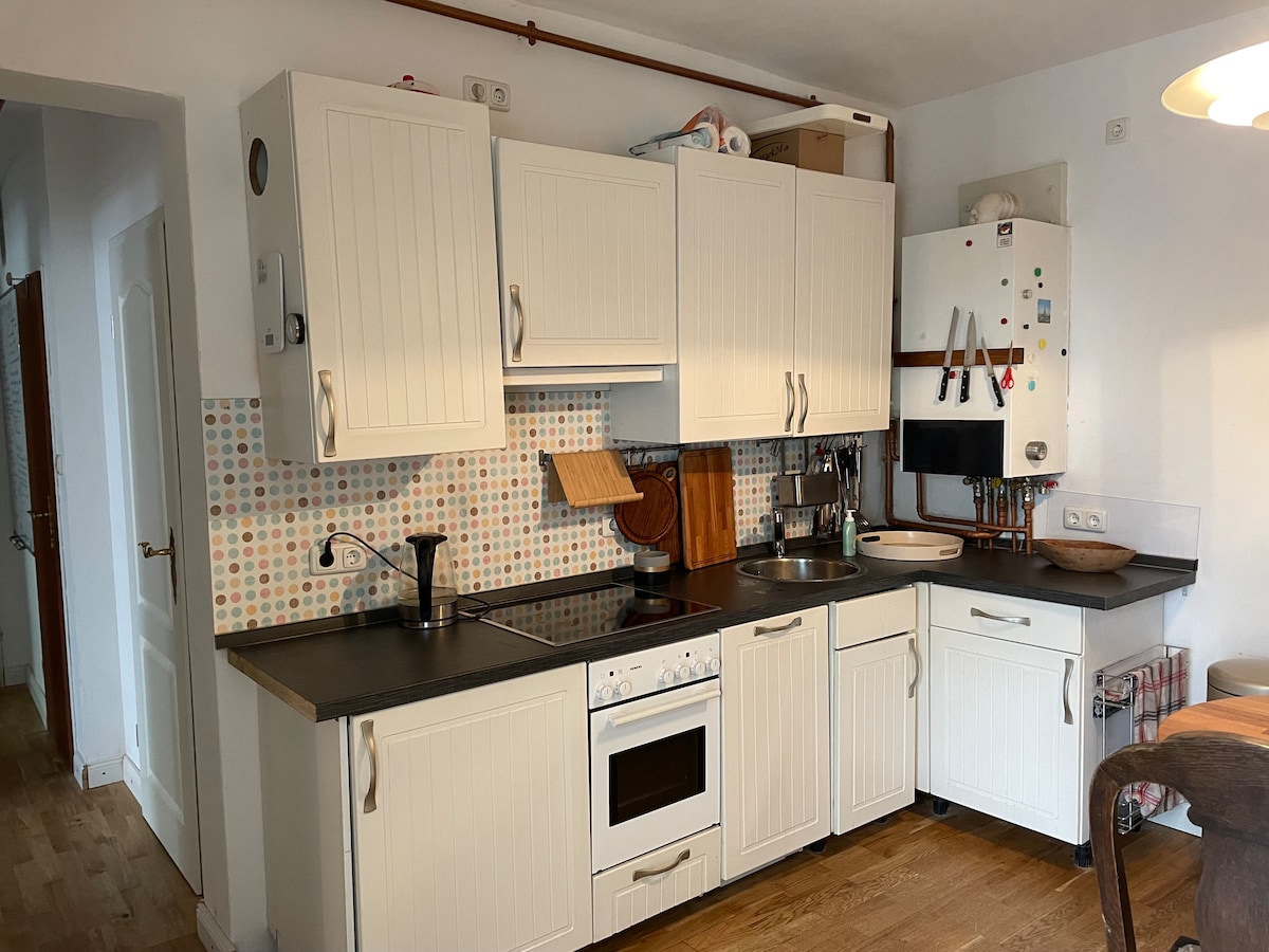 The kitchen is equipped with white cabinetry and a mix of modern and functional appliances. A tiled backsplash features a colorful pattern. Counter space is available for meal preparation, with a stovetop, oven, and a dishwasher integrated into the design. Wooden flooring adds warmth to the space.
