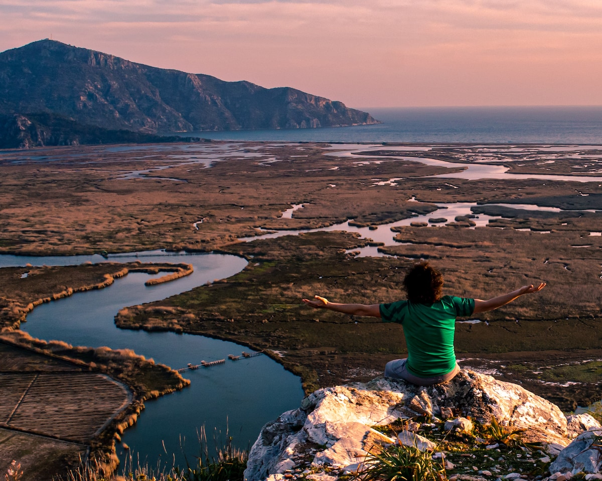 A person sits on a rocky ledge, arms extended, overlooking a vast landscape. The scene features winding rivers meandering through fields, with distant mountains rising in the background. A soft orange hue from the setting sun casts a warm glow across the land and water.