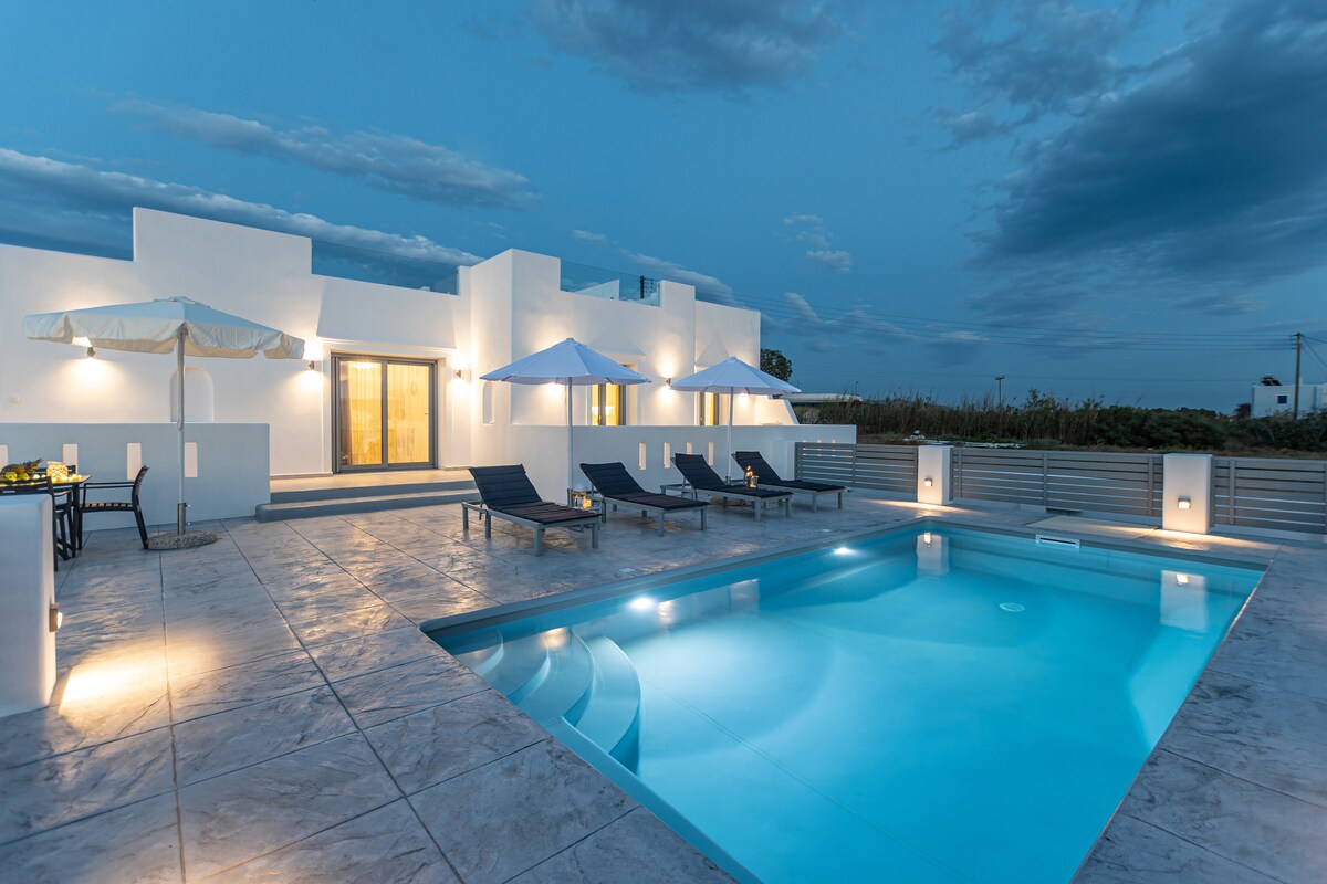 An outdoor area is illuminated by soft lighting, showcasing a clear swimming pool and several lounge chairs. Two umbrellas provide shade next to the pool. The villa's facade features large glass doors, enhancing the serene evening ambiance.