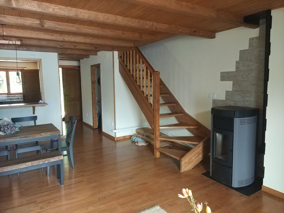 A spacious living area features wooden beams and a warm wooden floor. A staircase leads to the upper level, while a modern stove stands beside a large dining table that accommodates multiple guests. Natural light fills the room through visible windows.