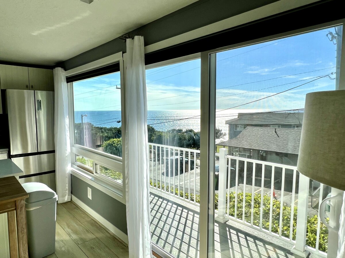 Large windows frame the serene ocean view, allowing natural light to fill the open living space. A glimpse of the neighboring home and a white railing can be seen outside, enhancing the connection between the interior and the coastal landscape.
