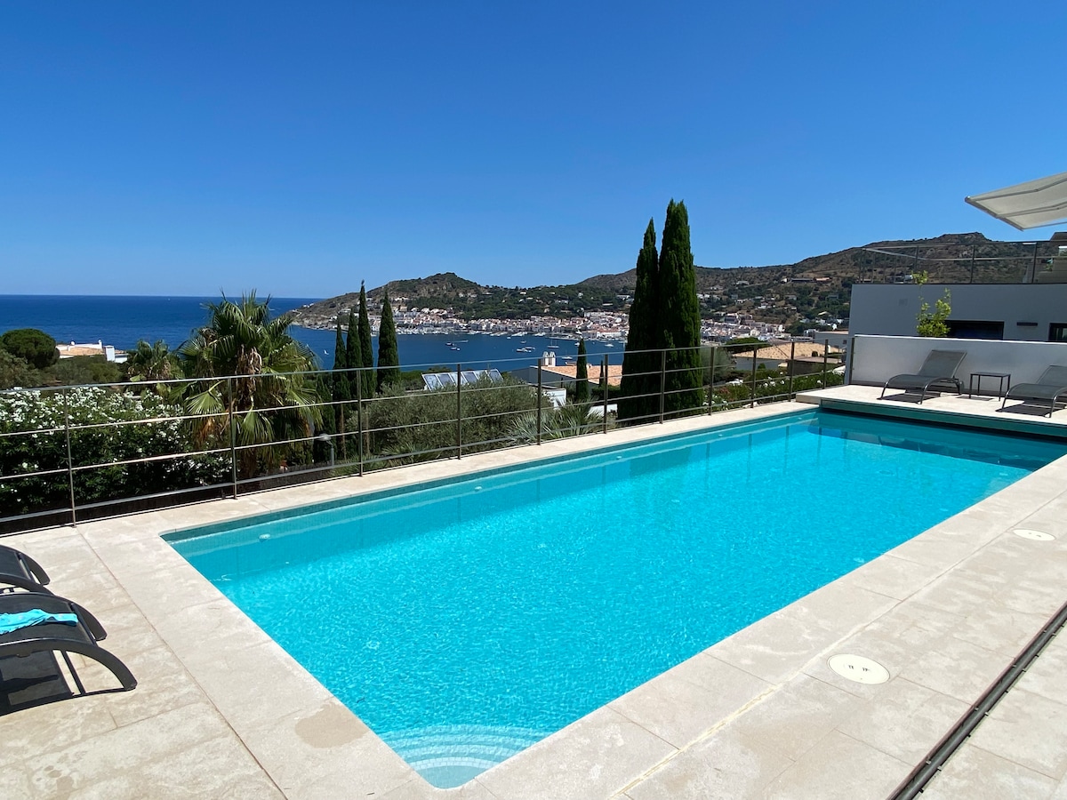 A large, inviting pool with clear blue water is surrounded by a spacious deck. Lush greenery and tall trees line the perimeter, while distant views of the village and coast are visible in the background under a clear blue sky.