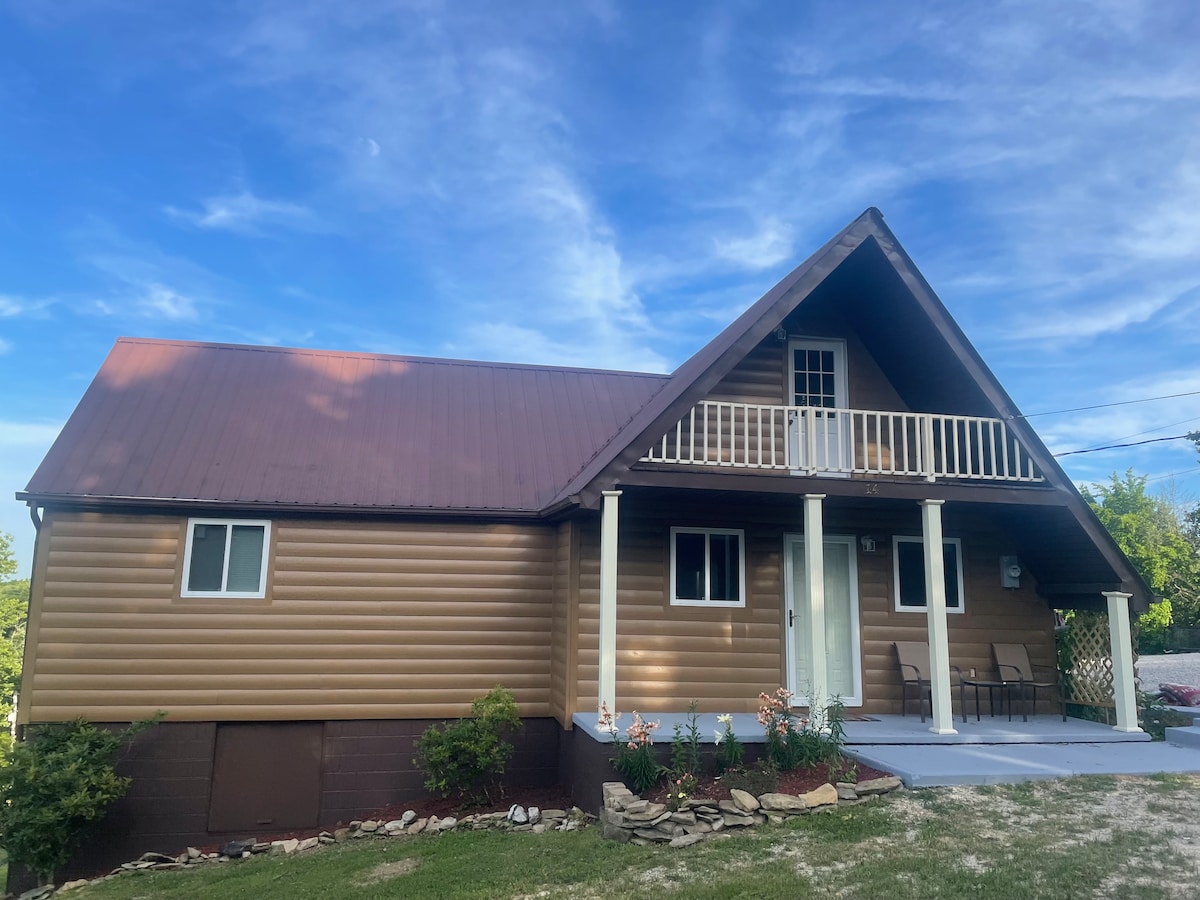 The cabin's exterior is showcased with a sloped roof and warm wood paneling. A covered porch features two chairs, with flowering plants and a well-kept lawn in the foreground. Large windows allow natural light, while the sky above adds an expansive feel to the setting.