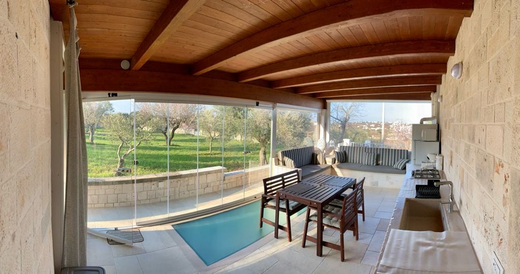 A covered outdoor area features a small pool and a wooden dining table surrounded by chairs. Glass walls provide views of the lush green landscape, with trees and an open field visible. The space is complemented by a seating area and kitchen appliances.