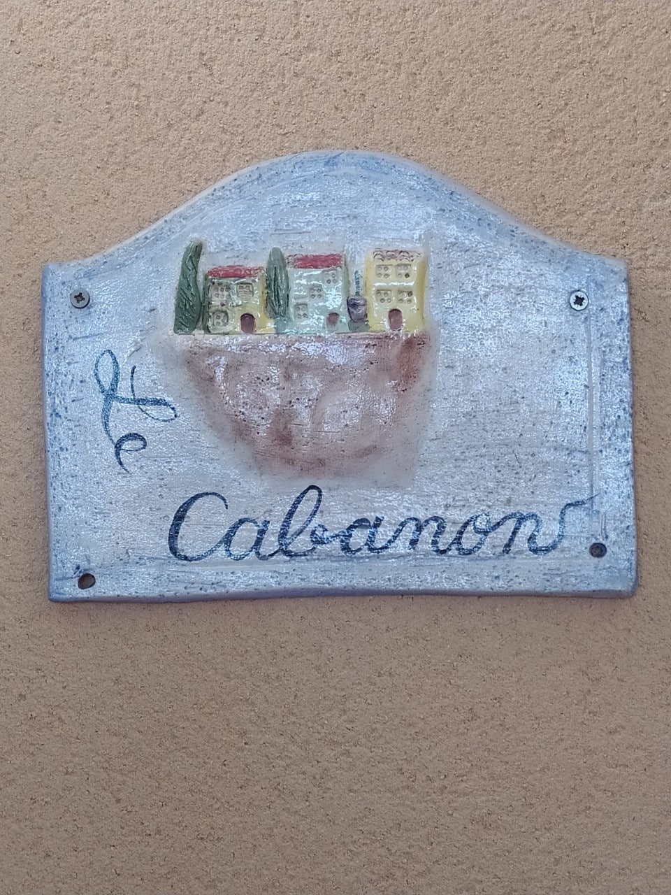 A rustic sign displays the name 'Le Cabanon' in a flowing script, accompanied by a colorful bas-relief depiction of small houses and trees. The sign is fixed to a textured, light-colored wall, adding character and local charm.