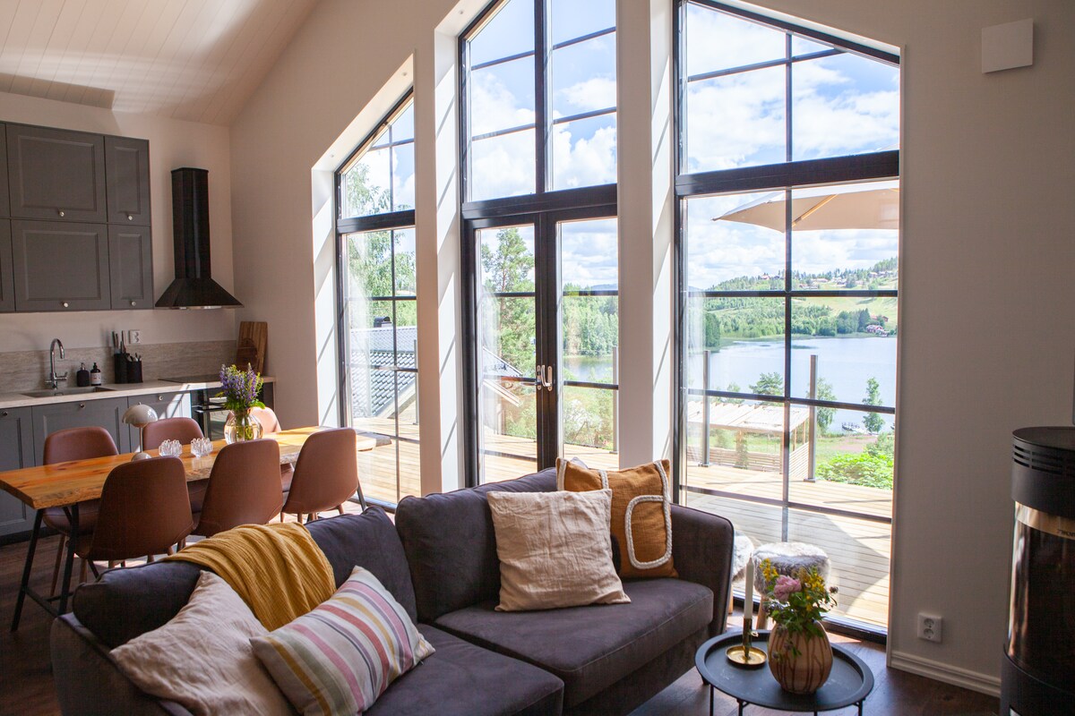 Large windows allow natural light to fill the living area, providing views of the lake and surrounding greenery. The space features a cozy sofa with decorative pillows, a dining table with chairs, and a fully equipped kitchen in the background.